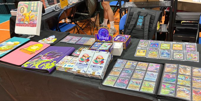 A vendor's table at a convention displays an array of colorful trading cards and accessories from the "vibes tcg" collection, along with merchandise featuring Pikachu, Mario, and Pokémon characters in pink and purple playmat sleeves. The spread aligns with the post, showcasing @vibes_tcg cards alongside Pudge Penguins and other gaming-themed products being sold and promoted.