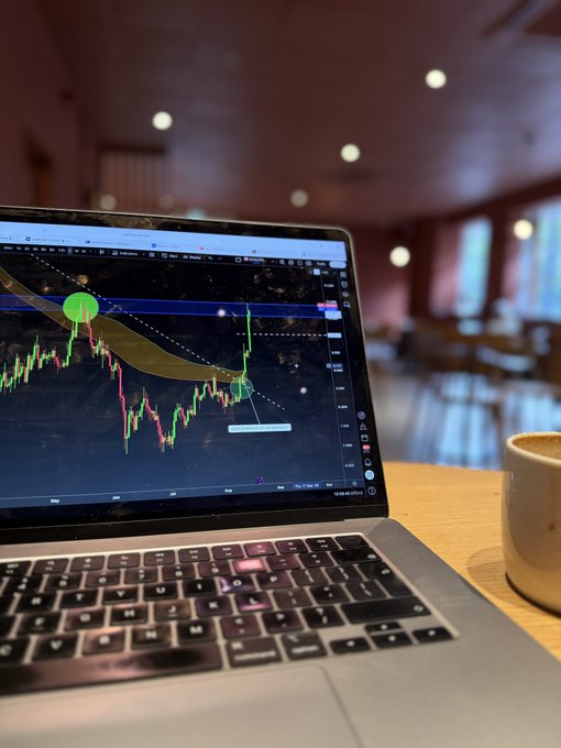 A laptop on a cafe table displays a candlestick chart with green and red price action and a green circle marker highlighting a recent upward move, accompanying the post's bullish announcement that the token has reclaimed the $10 level and a new video is coming.