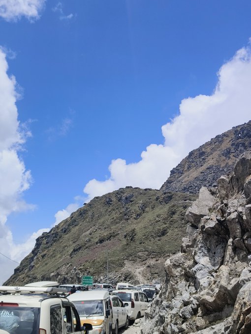 A massive traffic jam of vans and cars is backed up along a narrow mountain road beneath a steep rocky cliff, illustrating the 3-hour gridlock referenced in the post. The image perfectly captures the frustration of being stuck in a slow-moving line of vehicles, making the traffic option seem just as painful as a $300 trading loss.