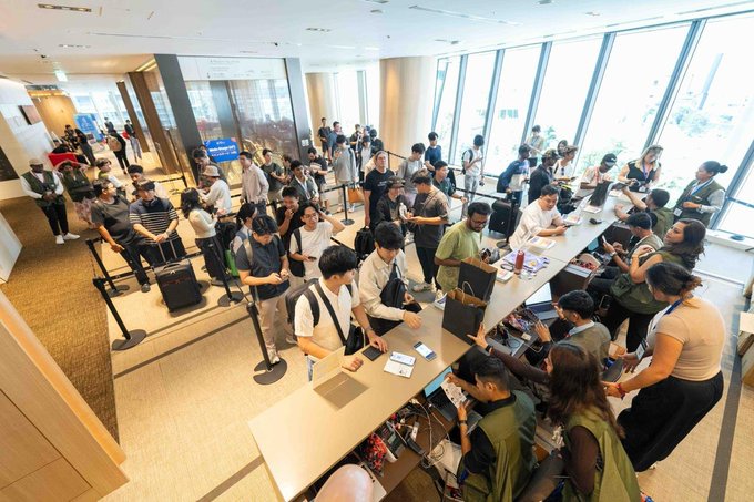 A crowded Apple Store showing long queues of customers being assisted by staff at long display tables filled with products during what appears to be a product launch event.