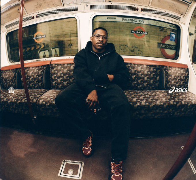 A man wearing glasses and a black hoodie sits on a patterned seat inside a London Underground train, with 'PADDINGTON' and 'NO SMOKING' signs visible in the window.