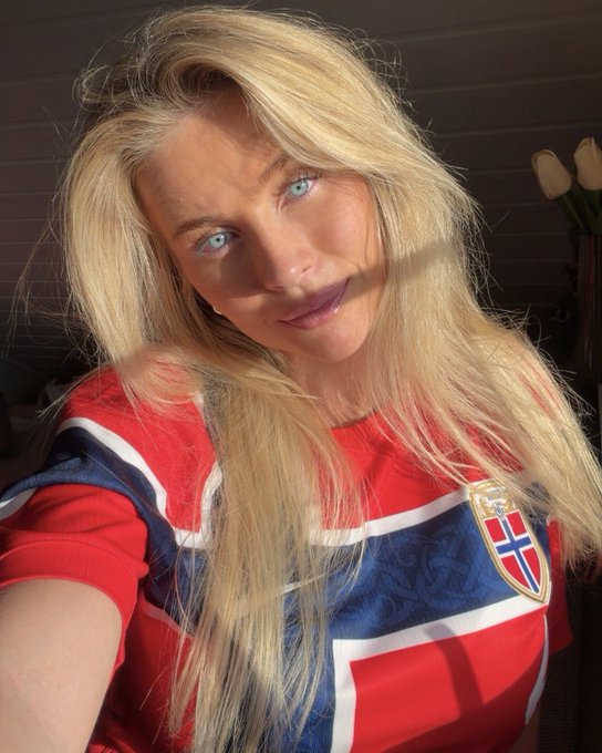 A selfie of a blonde woman wearing a red and blue Norway football jersey, taken indoors with warm lighting.