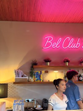 A neon pink "Bel Club" sign glows above a bar counter where two people are seated, with a woman in white smiling warmly while leaning close to a man in a blue polo shirt, capturing the lively social atmosphere of the Padel Rave Bangkok event.