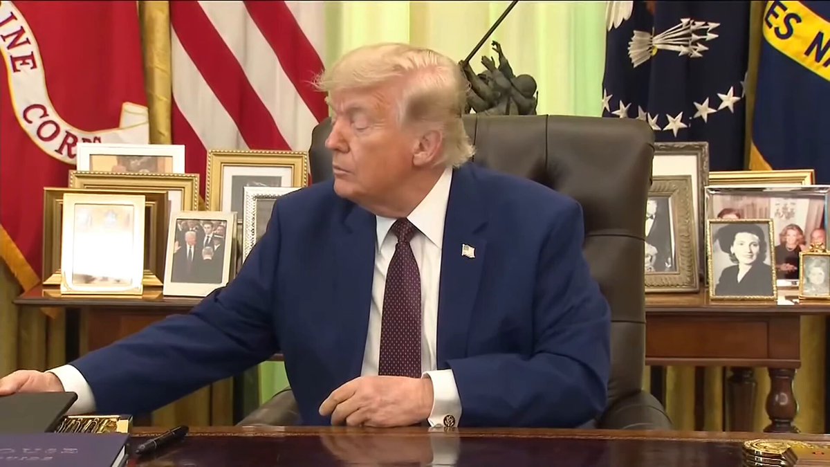 The image shows President Donald Trump sitting at his desk with a serious expression, as he signs an executive order. The Oval Office setting includes a chair, a desk, and several framed pictures hanging on the wall behind him. In addition to these elements, there are two people in the background who appear to be involved in some sort of discussion or meeting related to the President's actions.

The image also contains text that reads "BREAKING: President Trump signs major DOGE Executive Order, helping FIRE Deep State rogue federal workers in policymaking roles, and holding them accountable for undermining the America first agenda LFG! KEEP DOGEING". This suggests that there might be a political issue or controversy at hand involving the President's actions.