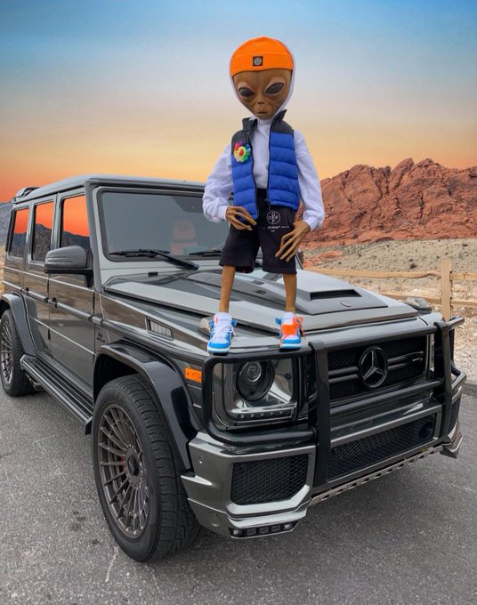 An AI-generated image featuring an alien character wearing a blue vest and orange beanie, standing on the hood of a black Mercedes-Benz G-Wagon against a desert sunset background.