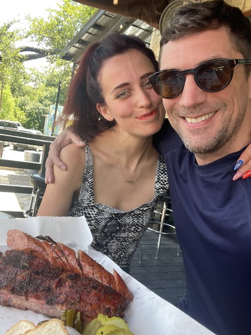 A selfie of a smiling couple at an outdoor restaurant with a plate of barbecued ribs and pickles visible in the foreground.