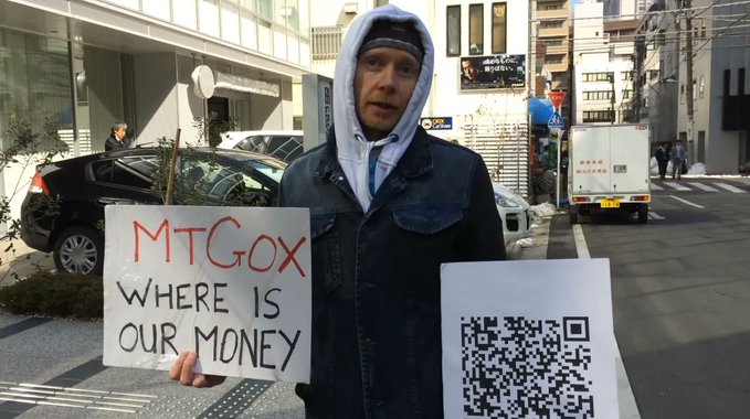 A man wearing a hooded jacket stands on a street holding a sign reading 'MtGox Where is our money' and another sign with a QR code, protesting outside.