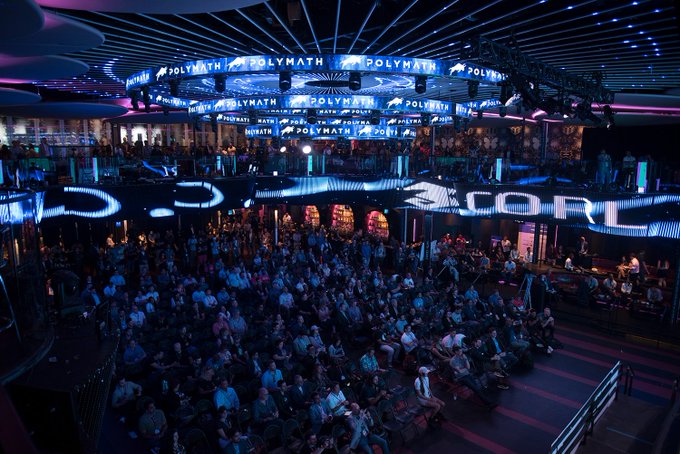 A large crowd fills an ornate venue illuminated with neon blue and purple lighting, where digital displays prominently feature the "POLYMATH" name along the upper balcony and a "CORE" logo on the lower level. The scene captures the Polymath Network's sponsorship presence at a major conference, visually tying into the announcement of their upcoming panel on the $16 trillion tokenization migration happening July 21 at 5 PM.
