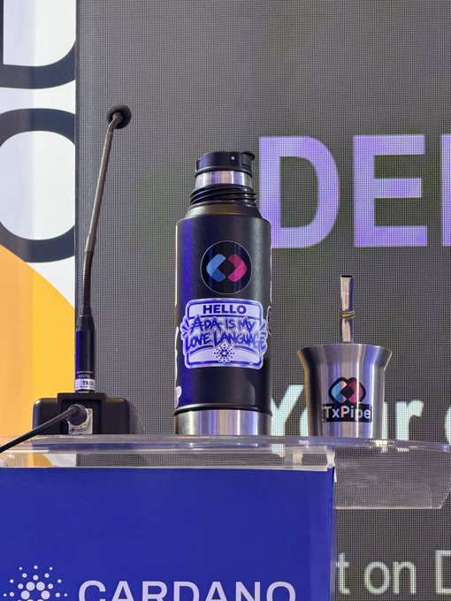 A black water bottle with a handwritten "HELLO ADA IS MY LOVE LANGUAGE" label displayed next to a metal cup with the TxPipe logo at a Cardano event.