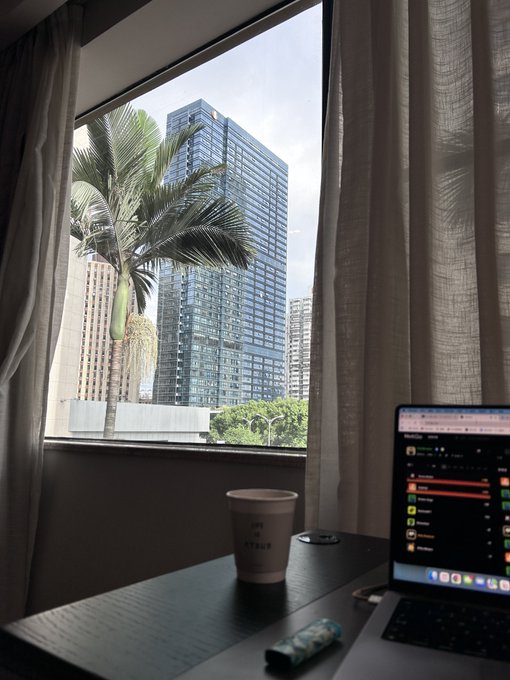 An indoor scene showing a desk with a laptop, a coffee cup, and a portable speaker, with a window view of a palm tree and a tall glass skyscraper against a cloudy sky.