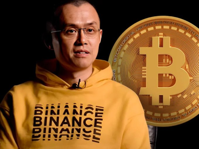 A man wearing a yellow Binance hoodie stands in front of a black background with a large golden Bitcoin graphic displayed beside him.