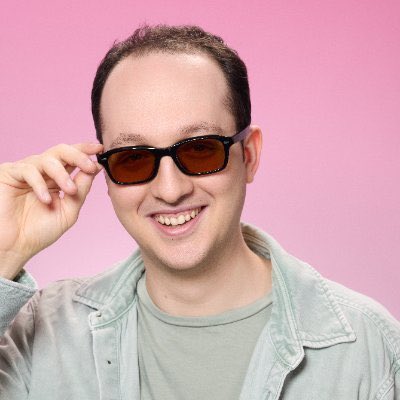 A man with short dark hair smiling and lowering a pair of dark sunglasses, wearing a light green shirt under a pale gray jacket, posed against a solid pink background.