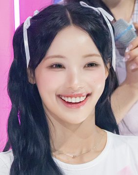 A close-up portrait of a smiling young woman with long dark hair styled in pigtails with white bows, wearing a white t-shirt.