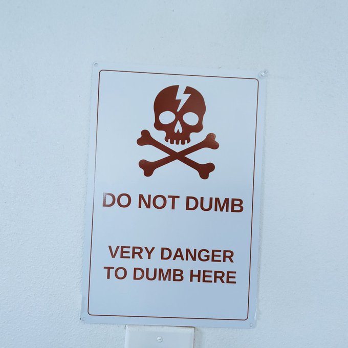 A white warning sign on a wall featuring a brown skull and crossbones icon, with text reading 'DO NOT DUMB' and 'VERY DANGER TO DUMB HERE'.