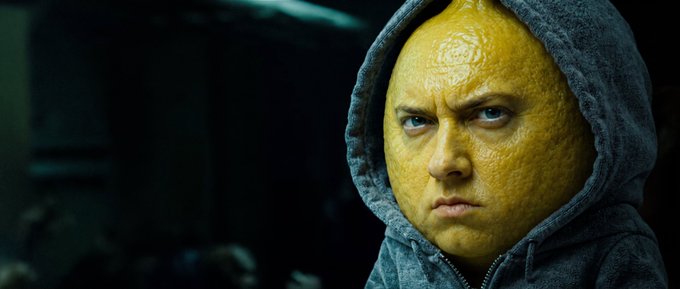 A hooded man wearing a grotesque, lemon-textured mask that covers the right side of his face glares intensely, his serious expression conveying determination and grit.

The image pairs perfectly with the post's "cooking" meme language—the lemon-masked face embodies the focused, slightly unhinged confidence of someone "locking in" a trade, with the sour citrus motif visually reinforcing the $lemonem ticker.