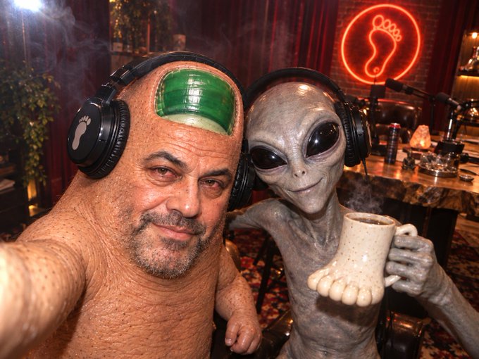 A bald man in alien makeup poses for a selfie alongside a gray alien, both wearing headphones in a dimly lit venue with a neon footprint sign. The image humorously captures a "beam me up" moment, with the alien offering a coffee mug shaped like a foot.