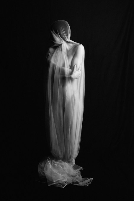 The image features a person standing against a black background, wearing a white dress or gown with a sheer veil draped over their head. The person's face is not visible due to the veils covering them. There are several pieces of text scattered around the image that read "Between Almost and Real" and "between almost real". These texts seem unrelated to the main subject of the photo, which is the person in the white dress or gown with a veil.