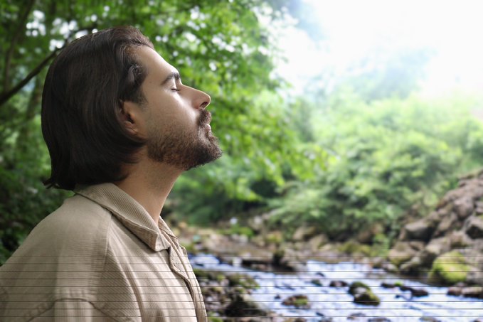 A man with long dark hair and a beard stands in profile with his eyes closed, peacefully breathing in fresh air beside a rocky stream in a lush green forest.