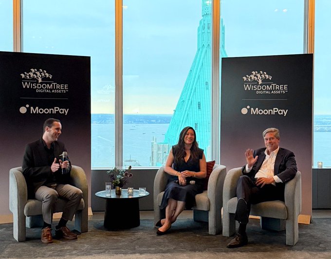 Three people sit in armchairs on a stage with "WisdomTree Digital Assets" and "MoonPay" banners behind them, with a large window revealing a cityscape and water view. The post celebrates "Caroline D. Pham Day," likely referencing one of the panelists pictured.