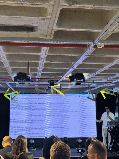 A man in a white shirt stands on a stage presenting in front of a large screen displaying text, while several audience members watch from the foreground under an industrial-looking ceiling.