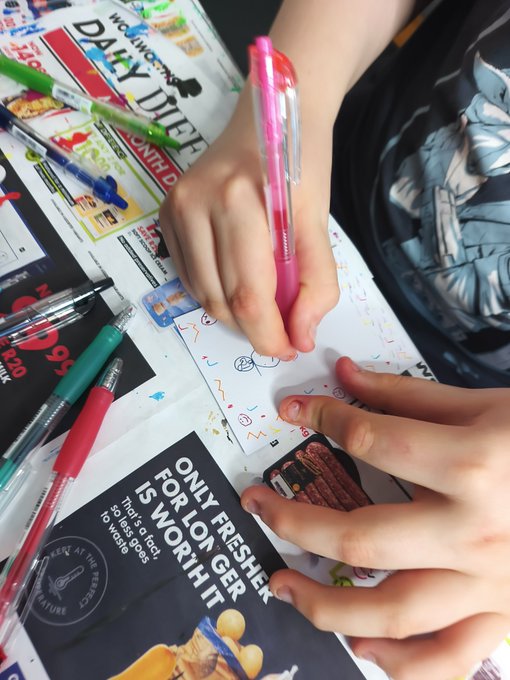 Close-up of hands writing with a pink pen on a card over various magazines, with visible text including 'DAILY DIET' and 'ONLY FRESHER... FOR LONGER IS WORTH IT'.