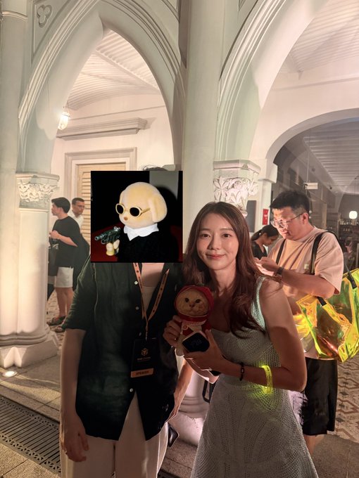 Two attendees pose together at the BNBChain SG event, with the woman on the right holding a Labubu keychain and the person on the left (face obscured) standing next to a prominent column; the post highlights the lively atmosphere and tags @jadoodoo_ at the @fourdotmemezh booth.