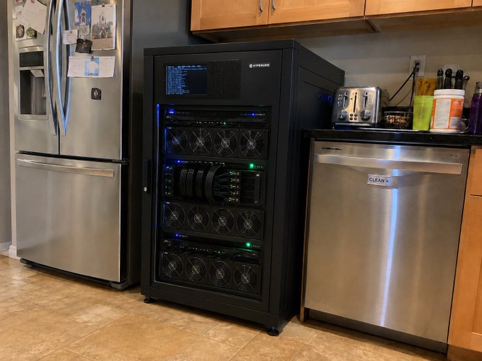 A home server rack with multiple hard drive bays and a small display sits in a kitchen between a Samsung refrigerator and a stainless steel dishwasher, illustrating the DIY infrastructure required for self-custody of data—a stark contrast to the convenience of relying on tech giants like Apple, Google, or Amazon.