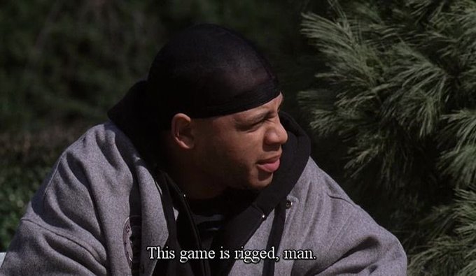 A screenshot from a show featuring a man in a black durag and gray hoodie outdoors, with the subtitle 'This game is rigged, man.'