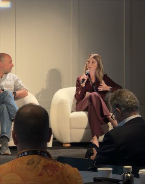 A panel discussion scene where a woman in a burgundy suit speaks into a microphone while seated in a white chair, with other attendees listening around her.
