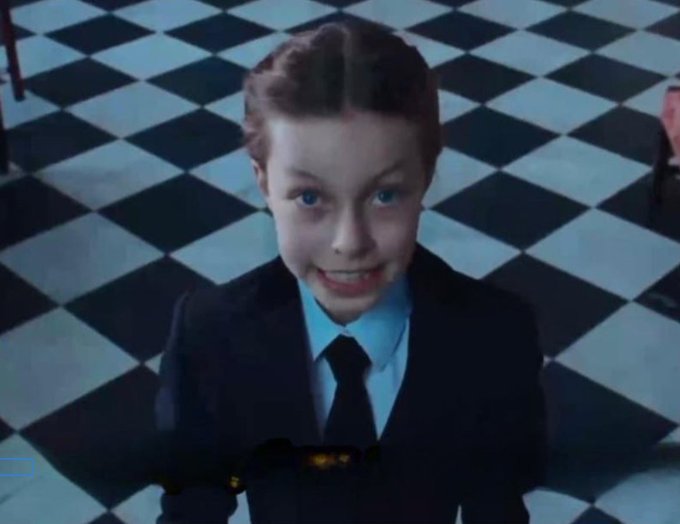 The image shows a young girl wearing a suit, tie, and blue shirt with her eyes closed as if she's smiling or making a funny face for the camera. The background features a black and white checkered floor that adds an interesting visual element to the scene. There is also text in the image that reads "now i want GLM 5.2 and Minimax M3".