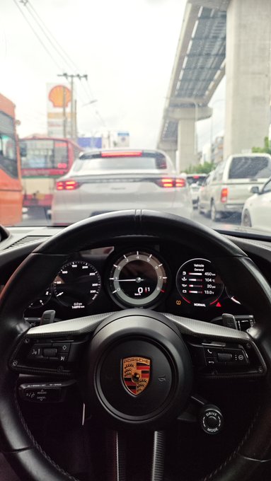 A Porsche driver's perspective from behind the steering wheel, stopped in traffic under an elevated highway. The digital dashboard shows the car in 2nd gear at 0 mph, with a speedometer reading and various vehicle stats including "13.5" visible, while the road ahead reveals stopped traffic including a Shell gas station and an Audi — capturing the everyday frustration of "Evening lads ♘" stuck in commute gridlock.