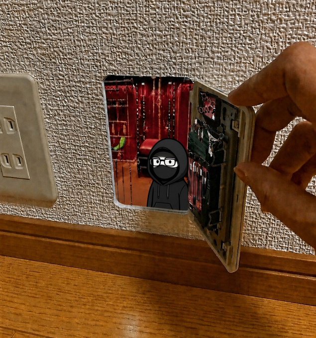 A hand opens a small decorative door mounted on a wall near an outlet, revealing a cartoon figure in a black hoodie inside a red-lit room.