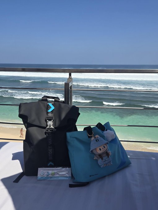 A black "BITGET WALLET" branded backpack and a blue tote bag with a cute plush keychain sit on a table overlooking a stunning ocean view at Coinfest Asia, likely belonging to a team member there to share exclusive merchandise during interviews.