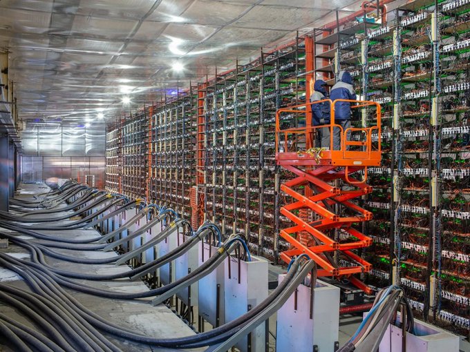 A worker in blue overalls stands on an orange scissor lift inside a large cryptocurrency mining facility, surrounded by rows of mining rigs with cooling fans and extensive power cables running along the floor.