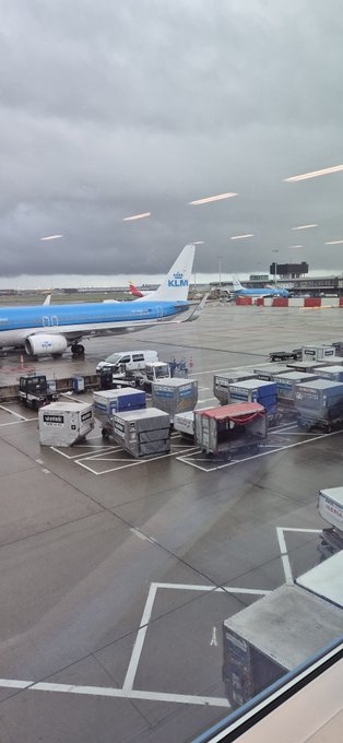 A view from an airport terminal window showing a KLM airplane at the gate with various ground service equipment, cargo containers, and baggage carts on the tarmac under a cloudy sky.