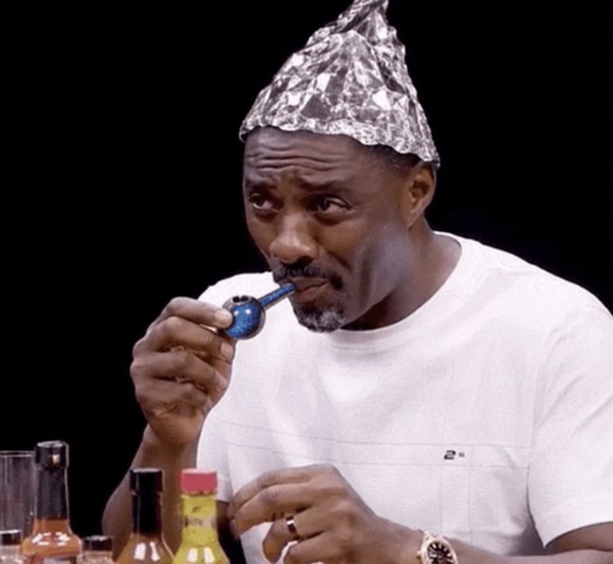 A man wearing a white t-shirt and a tin foil hat thoughtfully examines a blue glass pipe, with several hot sauce bottles visible in the foreground.