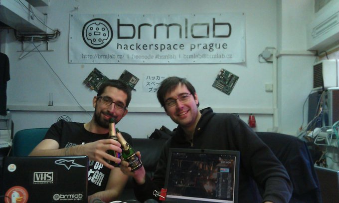 Two men, identified as "Slush" and "Stick," celebrate the 12th anniversary of founding Trezor at brmlab hackerspace Prague, toasting with drinks in front of laptops and a banner displaying the brmlab logo.