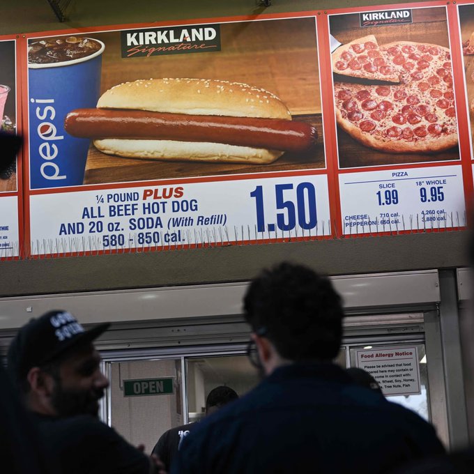 A Costco food court menu board displaying a Kirkland Signature ¼ Pound Plus All Beef Hot Dog and 20 oz soda for $1.50, alongside pizza prices.