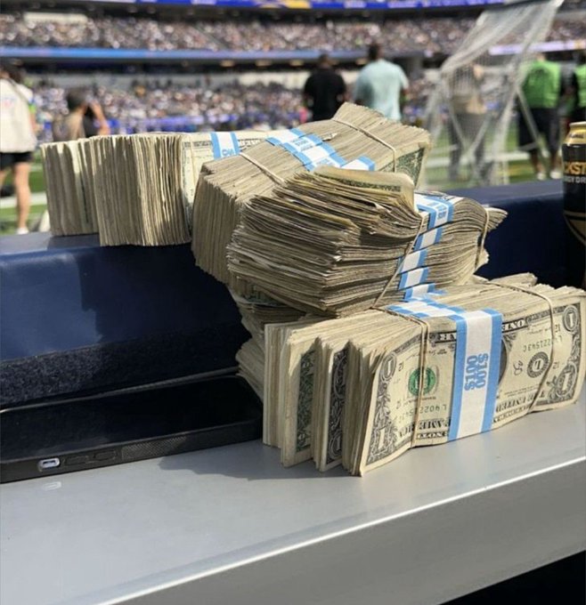 Stacks of cash bundles banded with blue straps rest on the railing of a packed stadium, suggesting a promotional giveaway offering the first 500 likers a chance to win unexpected money.