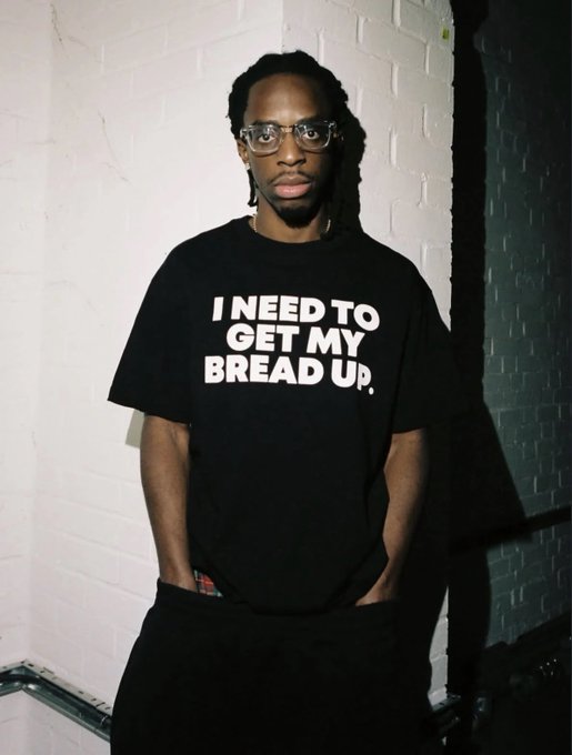 A young man with glasses stands against a white brick wall wearing a black T-shirt that reads "I NEED TO GET MY BREAD UP," channeling a motivational hustle vibe that fits perfectly with the post's upbeat "gm" and "let's win" energy to start the week.
