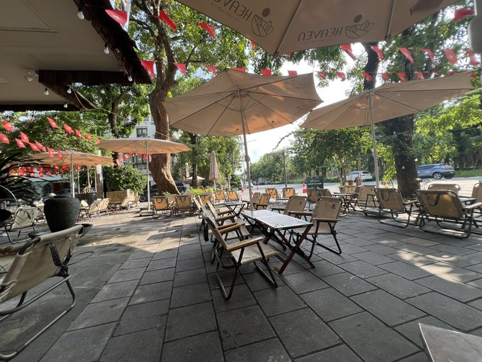 An outdoor restaurant patio features beige Heineken-branded umbrellas, red pennant flags strung between trees, and rows of empty folding chairs and tables on a tiled floor.