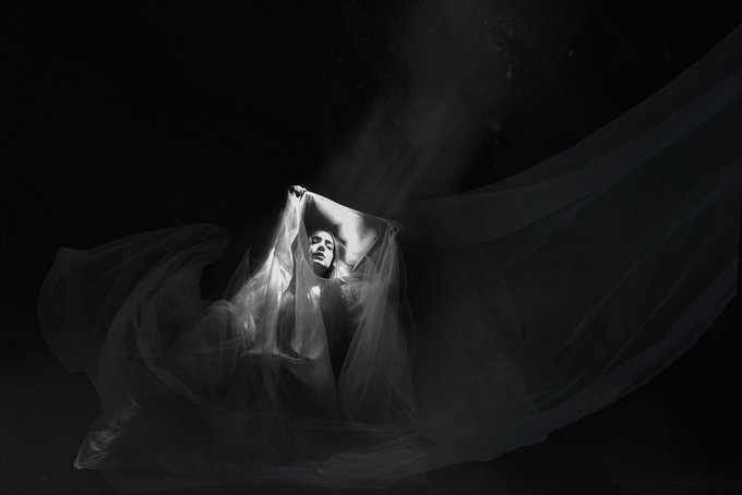 The image features a woman with her arms raised, holding a white sheet or veil that appears to be floating in front of her face. The background is dark, which contrasts with the light source shining down from above, creating an ethereal atmosphere. The photo has been edited and posted on social media platforms like Instagram, where it has received numerous likes and shares.