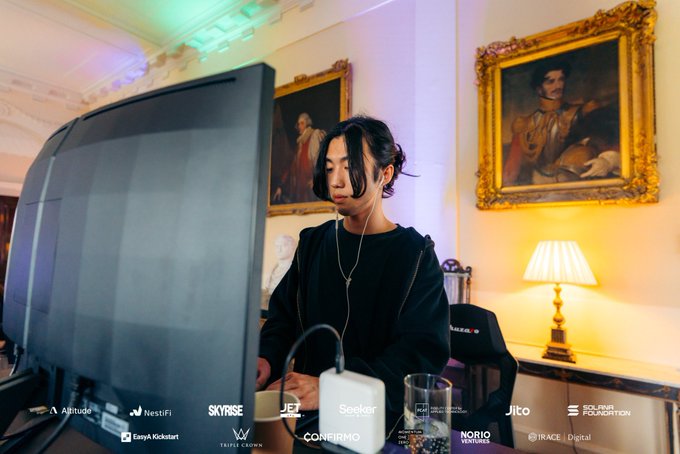 A young man with dark hair wearing a black sweatshirt working at a computer monitor in an elegant room with classical paintings and a lamp. The image features various sponsor logos at the bottom including Altitude, NestFi, Skyrise, Jet, Seeker, Coinbase, Norio Ventures, Jito, Solana Foundation, I race digital, Triple Crown, and Easya Kickstart.