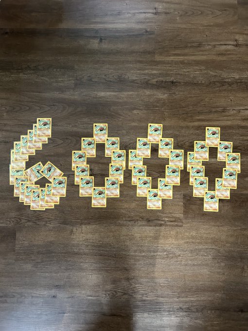 Arranged on a wooden floor, dozens of Pokémon Kabuto cards are artfully laid out to form the number "6000," celebrating the milestone of reaching 6000 Kabutos in the collector's epic quest to crown the once-forgotten underdog the "King" of collecting.