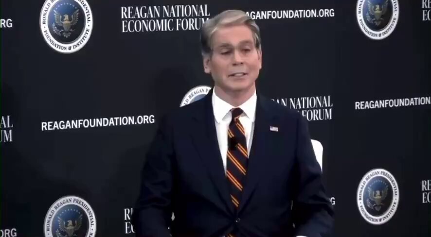 The image features a man dressed in a suit and tie standing against a black background with white text, including logos such as "Reagan National Economic Forum" and "Reagan Foundation". The man appears to be speaking or addressing an audience at an event.