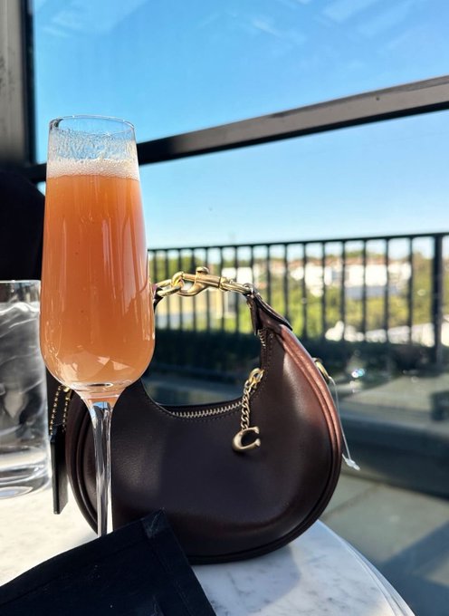 The image features a glass of orange juice sitting next to a brown purse on a table. The purse has a gold chain attached to it. The background shows a view of buildings, suggesting that they are located near an urban area or cityscape.