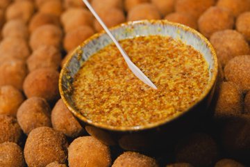 A close-up food photo of a bowl of reddish dip or sauce with a spoon, surrounded by breaded fried balls.