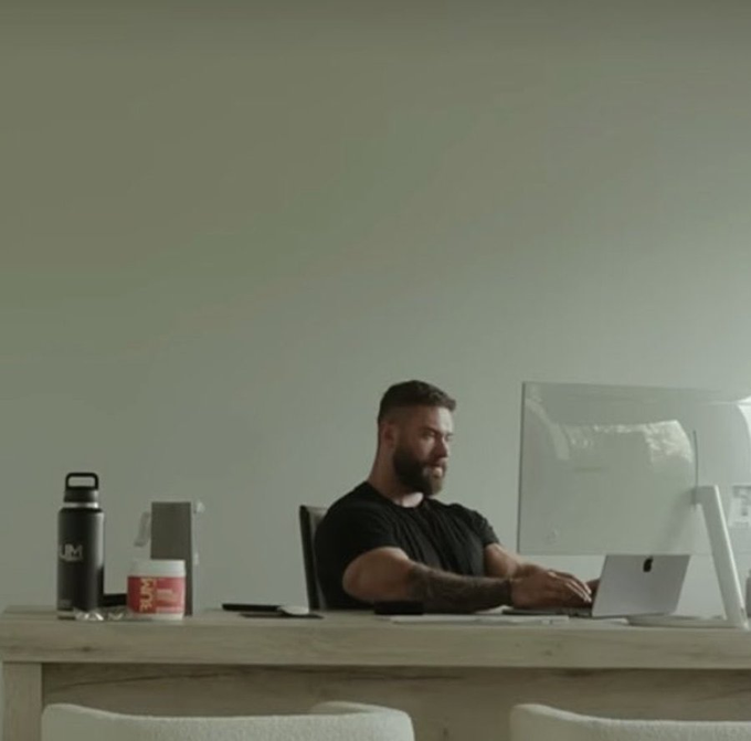 A bearded man in a black t-shirt sits at a wooden desk typing on a laptop next to a larger monitor, with a black water bottle, phone, and supplement tub nearby, visually reinforcing the post's casual lifestyle angle for "Chad" promoting Card Outpost as a trading card platform.