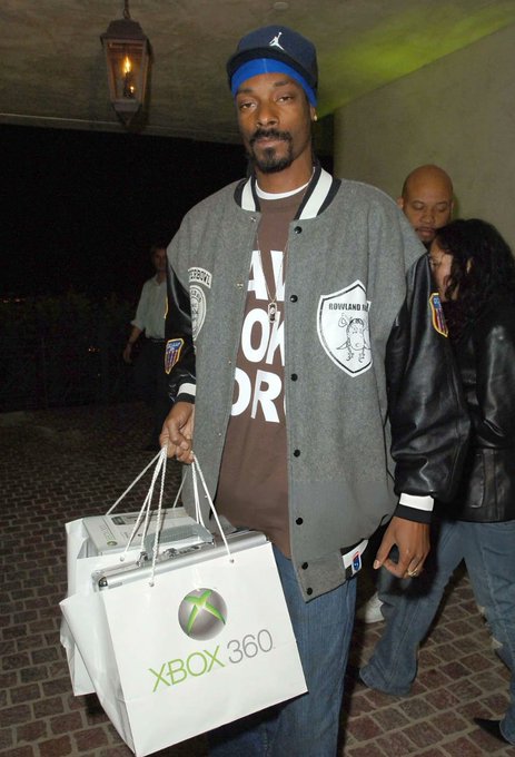 A man in a gray varsity jacket, blue beanie, and brown t-shirt carrying multiple white shopping bags branded with the Xbox 360 logo.