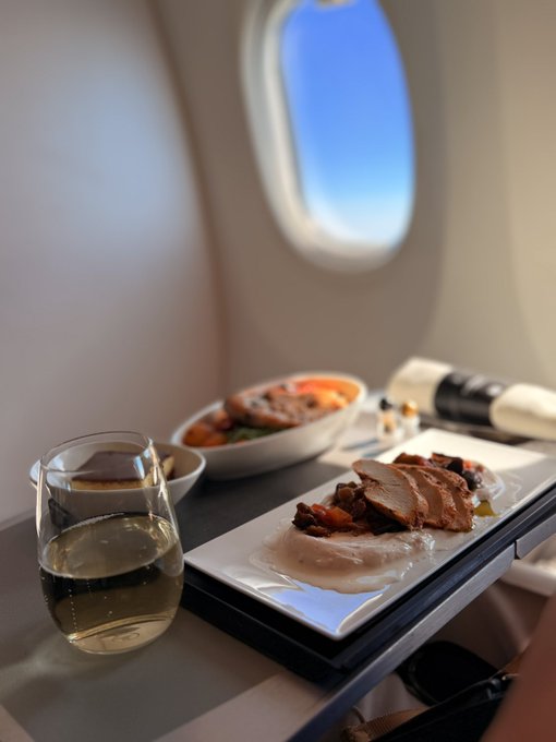 A glass of white wine accompanies a plated meal of sliced meat and a side dish, served on a tray by an airplane window, fitting the "back to sweet home" travel-themed post about returning home.